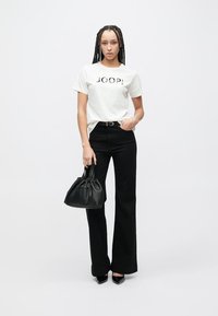 Camiseta blanca con texto "JOOP!", pantalones negros acampanados de talle alto, bolso negro y tacones puntiagudos negros. Textura lisa, diseño casual.