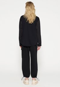 Zwarte top met lange, doorschijnende kanten mouwen, gecombineerd met losse zwarte broeken met verstelbare manchetten en een zijzak. Lichtgekleurde schoenen.