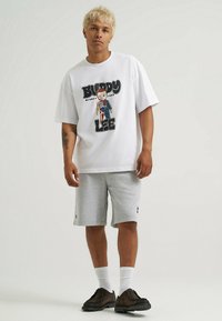 Biała koszulka graficzna z nadrukiem kreskówkowym oraz tekstem, w połączeniu z szarymi szortami, białymi skarpetkami i ciemnymi butami. Gładkie tło.