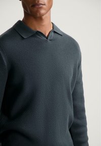 Man wearing a dark blue textured knit sweater with a collar against a plain light background, showing upper torso and neck.
