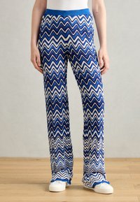 Blue and white zigzag-patterned knit pants with a blue elastic waistband, featuring a relaxed fit and textured fabric. White sneakers are worn.