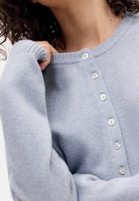 Ljusblå stickad kofta med rundad halsringning och stora, pärlemorskimrande knappar längs framsidan; mjuk textur, ribbade muddar.