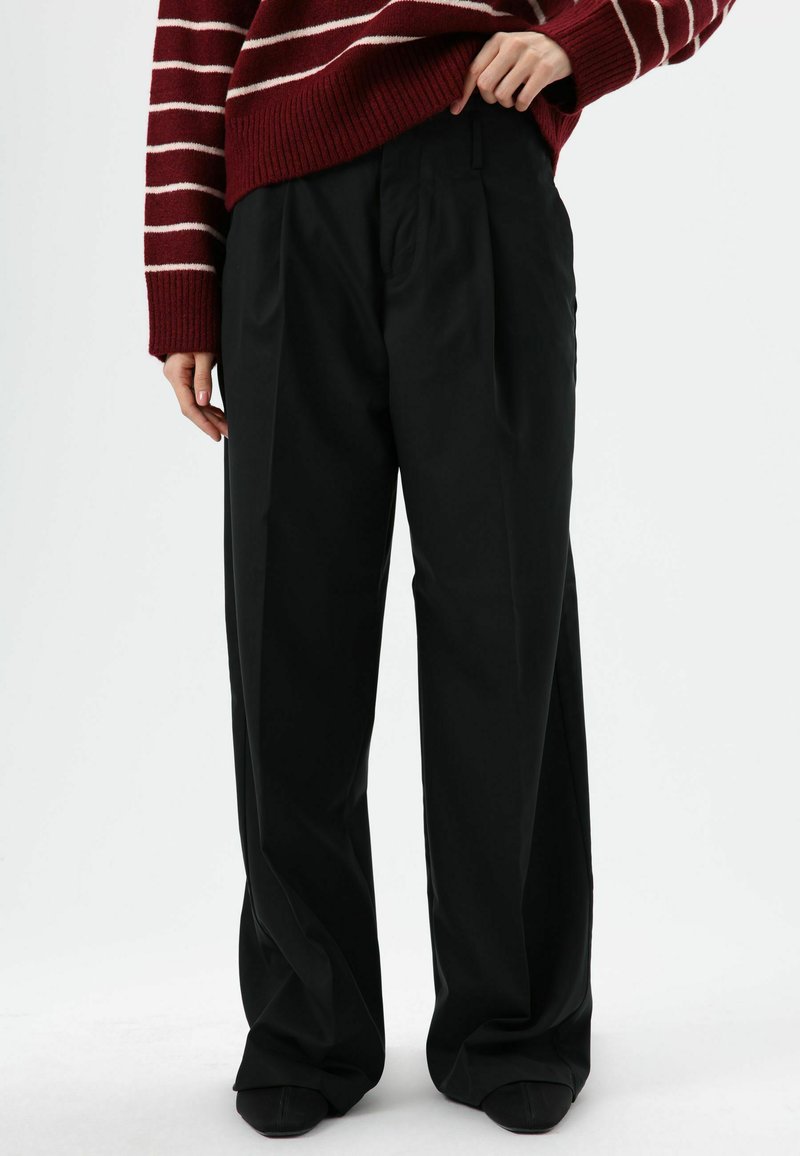 Zwarte broek met wijde pijpen en plooien aan de voorkant, gemaakt van gladde stof. Draagt een bordeauxrode en crèmekleurige gestreepte gebreide trui.