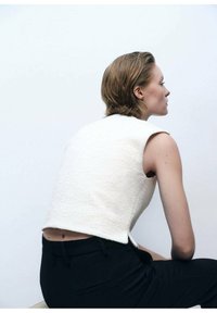 Weißes, strukturiertes schulterfreies Oberteil mit einem leicht verkürzten Design, kombiniert mit schwarzen Hosen. Fokus auf die einfache Silhouette und klaren Linien.