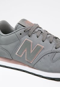 Baskets New Balance grises avec des accents roses, des lacets épais, un col rembourré et une semelle texturée, montrées en gros plan de profil.