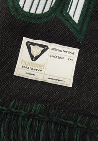 Etiqueta da marca desportiva Hummel em tecido preto com franjas verdes. A etiqueta inclui um logótipo, "Serving the Game" e detalhes de marca registada.