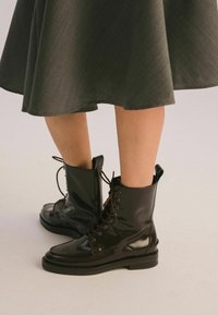 Czarne lakierowane botki za kostkę z okrągłymi czubkami, sznurowane z przodu, z brązowymi sznurówkami i masywną czarną podeszwą. Ustawione na tle szarej spódnicy.