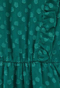 Zeleni materijal s teksturiranim točkicama, nabrani pojas i naborani vertikalni detalj na desnoj strani.