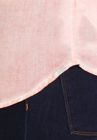 Chemise en lin rose clair avec un ourlet arrondi, présentant des bords cousus et une petite étiquette métallique, portée par-dessus un jean en denim foncé.
