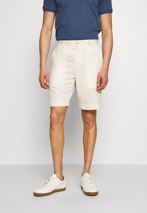 Hombre con pantalones cortos beige a medida, zapatillas blancas y una camisa de manga corta azul oscuro, de pie junto a una pared lisa.