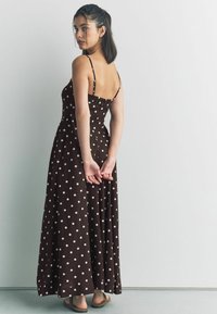 Brązowa maxi sukienka w kropki z spaghetti straps, dopasowaną górą i zwiewną spódnicą. Materiał jest lekki o gładkiej fakturze.