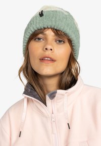 Žalia ir grietinėlės spalvos megzta skrybėlė su tekstūruotu paviršiumi ir juodu žymekliu. Dėvima su šviesiai rožine fliso striuke, turinčia pilką apykaklę.