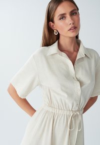 Šviesiai ruda lininė suknelė su apykakle, trumpomis rankovėmis ir raišteliu juosmenyje. Įrengtas mygtukų uždarymas ir švelni, lengva tekstūra.