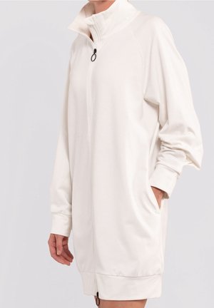 Femme portant une veste blanche à manches longues avec fermeture éclair, col montant et poches latérales, main reposant dans une poche.