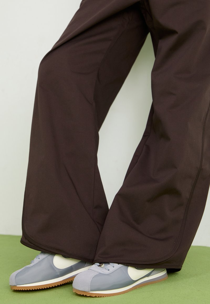 Braune weite Hosen mit glatter Textur und seitlichen Nähten, kombiniert mit grau-weißen Sneakers im Retro-Stil mit einer Gummisohle.