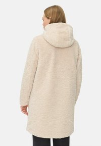 Cremfarbener, fusseliger Fleece-Mantel mit Kapuze. Hat eine lockere Passform, lange Ärmel und eine strukturierte Oberfläche. Der Saum ist leicht geschwungen.
