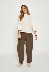 Krémový top s rukávmi ako pufy a diamantovým vzorom, kombinovaný s hnedými voľnými nohavicami a béžovými špicatými mulemi, na bielom pozadí.