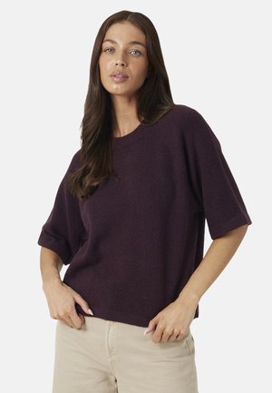 Femme aux longs cheveux bruns portant un pull violet foncé à manches courtes et un pantalon beige clair, posant avec les mains près de la taille.