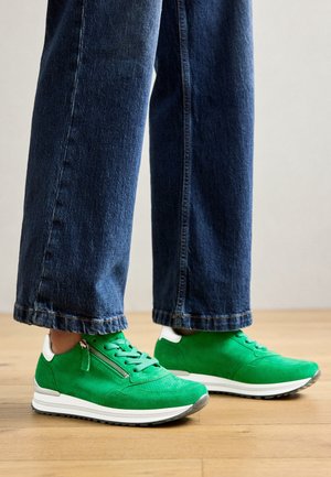 Person iført blå denimjeans og lysegrønne sneakers med hvide såler og lynlåse på siden, stående på trægulv.