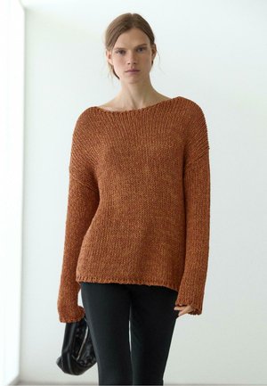 Femme aux cheveux châtain clair porte un pull en maille marron ample et un pantalon noir, tenant un sac noir sur un fond clair uni.