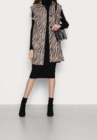 Beige en zwarte zebra-gestreepte vest met een kraaghals, schoudertabbladen, knoopsluiting en twee voorzakken, gecombineerd met een zwarte jurk en enkellaarsjes.