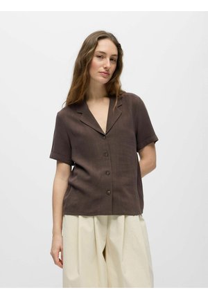 Femme aux cheveux longs et ondulés, portant une chemise marron à manches courtes avec des boutons et un pantalon ample couleur crème, debout devant un fond uni.
