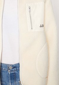 Kremowa kurtka polarowa z zamykaną na zamek kieszenią z białą tkaniną i szarym napisem. Miękka tekstura i luźny krój. Widoczny dżins w pasie.
