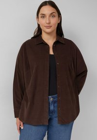 Bruine corduroy shirt met kraag en knoopsluiting, voorzien van een relaxte pasvorm en lange mouwen, gedragen over een zwarte top en blauwe jeans.