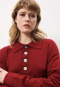 Pull en tricot rouge avec un col, ornée de boutons en argent et d'une texture douce. Le mannequin a les bras croisés et des cheveux châtains clairs.
