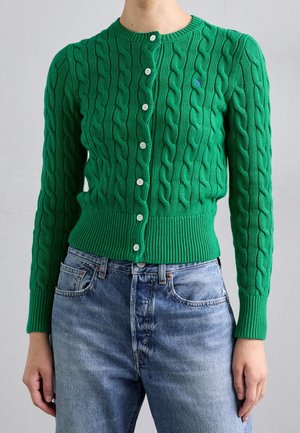 Cardigan vert en maille torsadée avec boutons et ourlet côtelé, porté avec un jean bleu taille haute sur une personne, sur fond uni.
