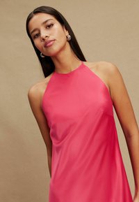 Rózsaszín, sima anyagból készült nyakbakötős ruha, folyékony szabással és minimális takarással. A modell arany karika fülbevalót visel, semleges háttér előtt.