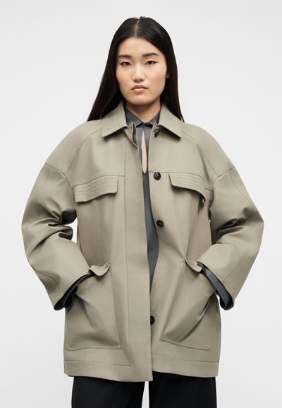 Νεαρή γυναίκα με μακριά σκούρα μαλλιά φοράει υπερμεγέθη μπεζ μπουφάν με τσέπες με καπάκι και μαύρα κουμπιά πάνω από σκούρο γκρι μπλουζάκι.