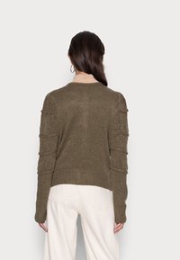Pull olive en vert avec un ourlet côtelé et des manches texturées à niveaux. Matière douce, col rond, porté sur un pantalon de couleur claire.