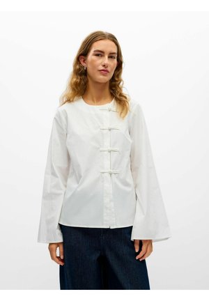 Femme aux longs cheveux ondulés portant un chemisier blanc à manches longues avec des nœuds sur le devant et un pantalon large bleu marine sur fond blanc.
