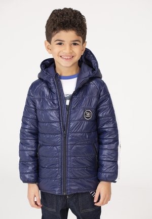 Smiling boy wearing a navy blue quilted jacket with hood and zipped front, standing against a plain white background.