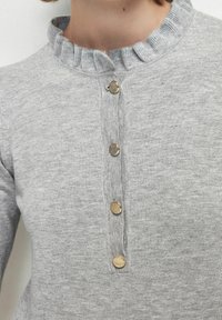 Pull léger gris avec un col à volants et une fermeture à boutons, doté de cinq boutons dorés sur le devant et d'une texture douce.