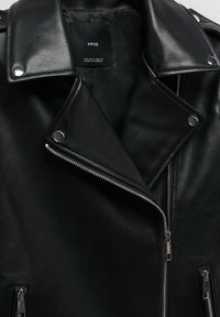 Veste en cuir noire avec fermetures éclair argentées et boutons-pression, avec un col plié et une étiquette de marque visible à l’intérieur.