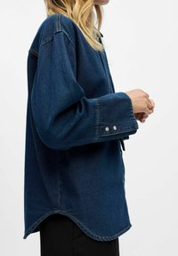 Camicia in denim blu scuro con bottoni, orlo curvo, vestibilità morbida e maniche lunghe. Presenta bottoni metallici ai polsini e dettagli di cuciture sottili.