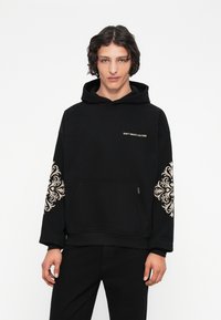 Melnā hoodie ar priekšējo kabatu un zelta paisley dizainu uz piedurknēm. Uz krūtīm ir zelta teksts "NEŠĶERĒ KULTŪRU".