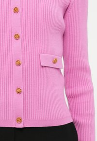 Nahaufnahme eines gerippten rosa Cardigans mit goldfarbenen dekorativen Knöpfen und einem kleinen Taschendetail auf der rechten Seite.