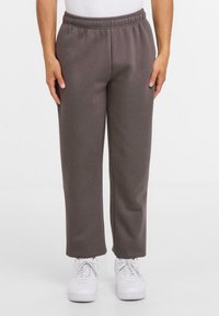 Person iført løstsiddende, mørkegrå sweatpants med elastisk talje og hvide sneakers på en ensfarvet hvid baggrund.