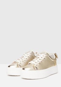 Goldene metallische Turnschuhe mit weißen Schnürsenkeln, runder Zehenpartie und dicker weißer Sohle. Verfügt über eine kleine Schleife an der Ferse. Glatte Textur durchgehend.