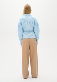 Light blue, collared jacket with gathered back design and voluminous sleeves, paired with loose, wide-legged beige trousers.