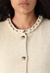 Cardigan beige avec un tricot texturé, présentant un col tressé et deux boutons ronds. Design simple avec un aspect doux et naturel.