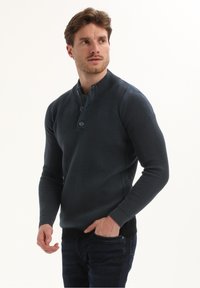 Man met lichtbruin haar, gekleed in een donkerblauwe gebreide trui met structuur en donkere spijkerbroek, kijkt opzij met een hand in zijn zak.