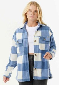 Blauw-wit geruite fleecejas met knoopsluiting, twee borstzakken en een kraag, gedragen over een wit topje en zwarte broek.