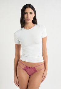 Giovane donna con capelli lunghi e scuri che indossa una maglietta bianca aderente a maniche corte e slip rosa in pizzo, su sfondo neutro.