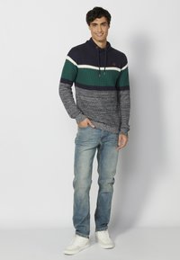 Sweter z dzianiny w niebieskie, zielone i białe paski z wysokim kołnierzem, z regulowanym sznurkiem i okrągłym detalem z logo. Noszony z jasnoniebieskimi dżinsami i białymi butami.