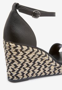 Sandalia de cuña de cuero negro con tiras al tobillo, que presenta un tacón de yute trenzado texturizado en un patrón de colores negro y natural.