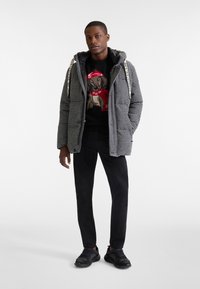 Sako s károvaným vzorom houndstooth, čierne a šedé, s kapucňou a šnúrkou. Pod ním čierny sveter s dizajnom psa. Tmavé nohavice a tenisky.